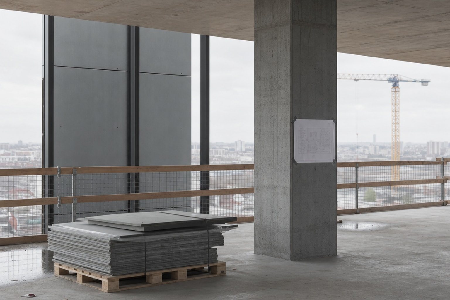 An upper floor during facade installation: grey panels stacked on a pallet, a drawing taped to a concrete column and a crane beyond the edge barrier.